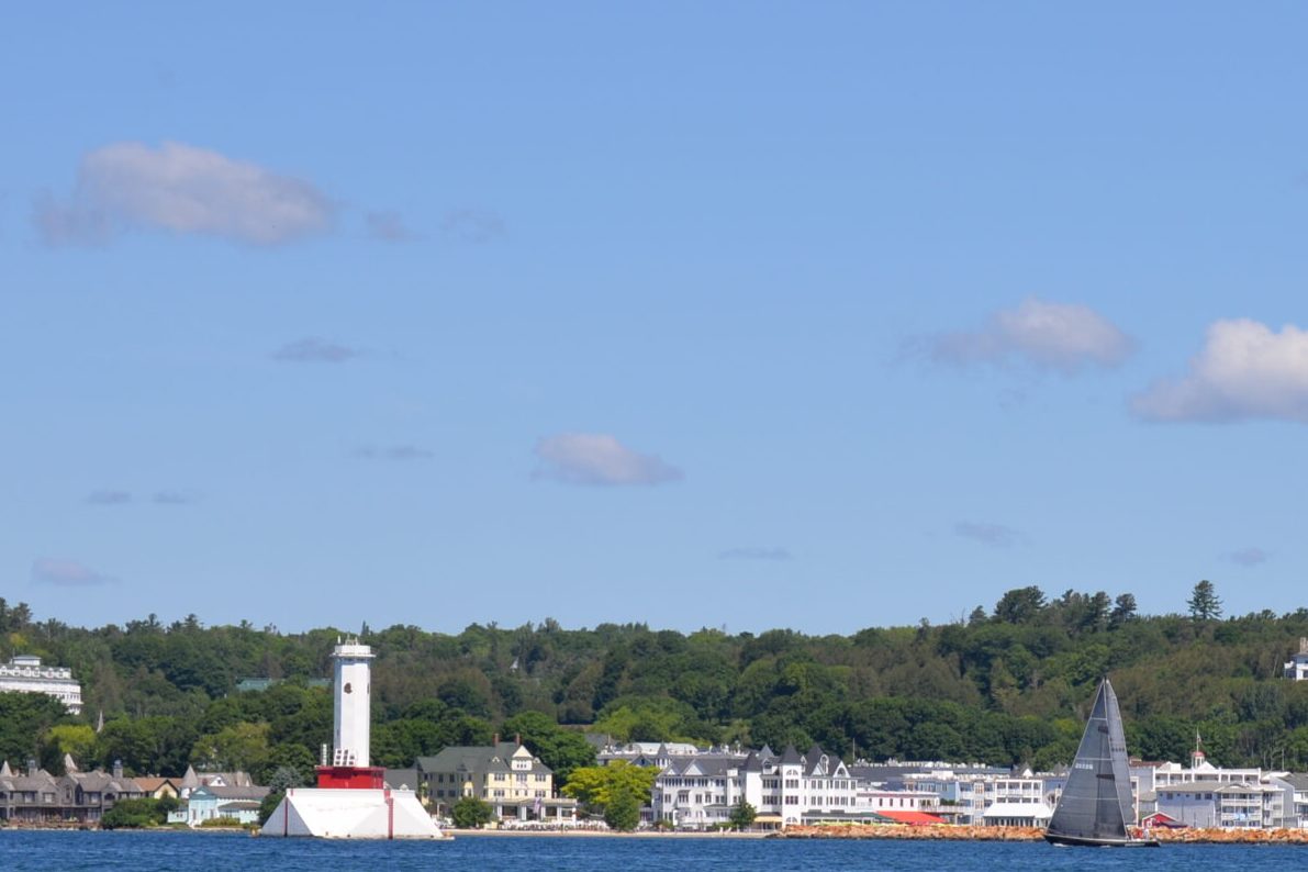 View of the Round Island Passage Lighthouse in the foreground, with the shoreline of Mackinac Island's business district visible in the background under a cloudy sky. This is the location where all sailing charters will begin.